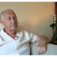 A person with short grey hair sits on a light-coloured sofa in a living room, wearing a white short-sleeved shirt and a wristwatch. In the background, a wooden shelf holds a potted plant and flowers.