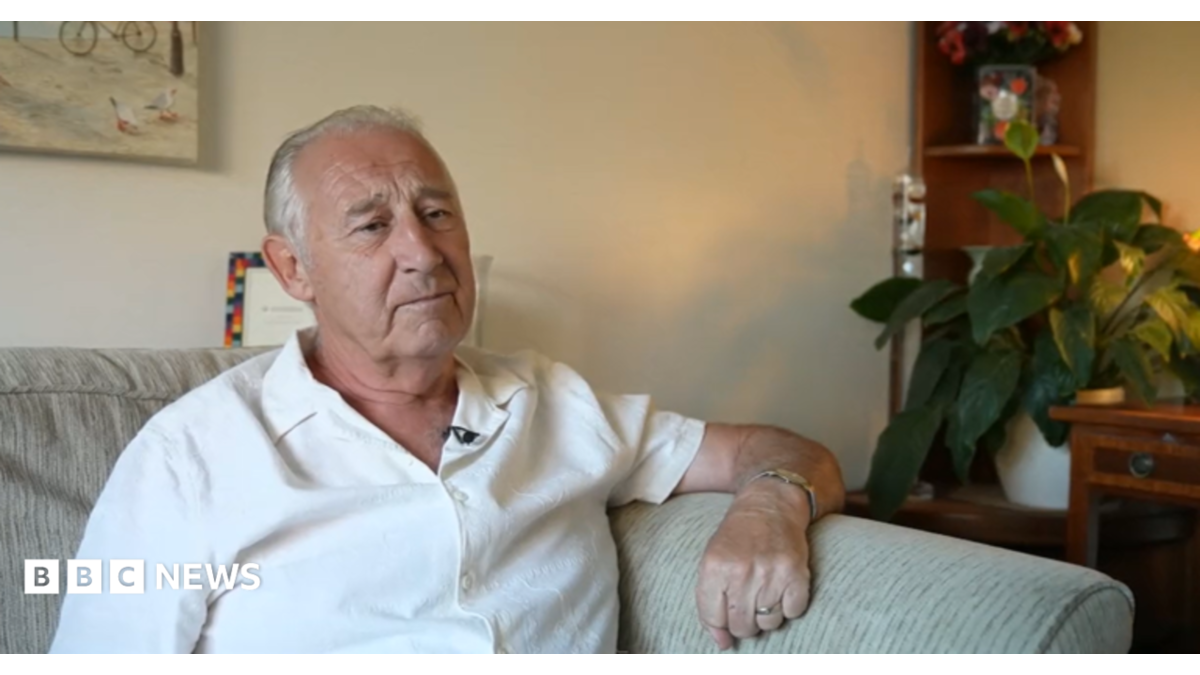 A person with short grey hair sits on a light-coloured sofa in a living room, wearing a white short-sleeved shirt and a wristwatch. In the background, a wooden shelf holds a potted plant and flowers.