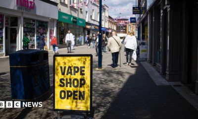 A high street scene in which there is a yellow sign with "Vape Shop Open" in black text. The sign is next to a bin. Pedestrians are in the background and there are shops either side of the walkway.