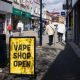 A high street scene in which there is a yellow sign with "Vape Shop Open" in black text. The sign is next to a bin. Pedestrians are in the background and there are shops either side of the walkway.