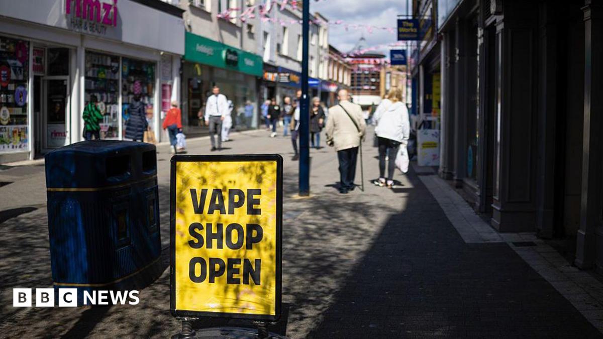 A high street scene in which there is a yellow sign with "Vape Shop Open" in black text. The sign is next to a bin. Pedestrians are in the background and there are shops either side of the walkway.
