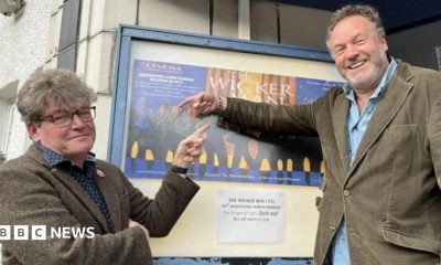 Two men in their 50s either side of a poster for The Wicker Man outside a cinema. Dominic, on the left, is in a tweed suit and has glasses and thick hair. Justin (right) is in a cord suit and has a beard. They are smiling and pointing at the poster.