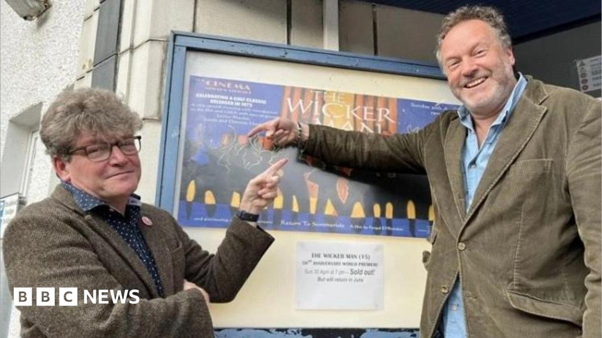 Two men in their 50s either side of a poster for The Wicker Man outside a cinema. Dominic, on the left, is in a tweed suit and has glasses and thick hair. Justin (right) is in a cord suit and has a beard. They are smiling and pointing at the poster.