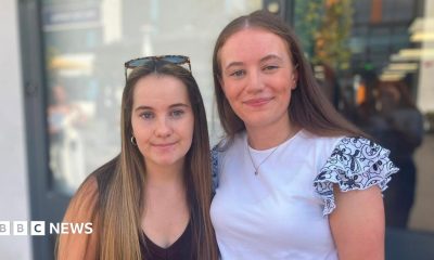 Beatrice and Amelia stand side by side outdoors, looking towards the camera. Amelia is wearing a dark sleeveless top with sunglasses resting on their head, while Beatrice is wearing a white top with patterned ruffled sleeves. The background is softly blurred and shows a modern building entrance.