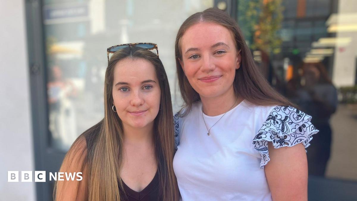 Beatrice and Amelia stand side by side outdoors, looking towards the camera. Amelia is wearing a dark sleeveless top with sunglasses resting on their head, while Beatrice is wearing a white top with patterned ruffled sleeves. The background is softly blurred and shows a modern building entrance.
