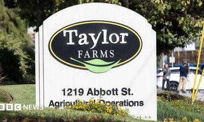 A curved white sign with a black sideways oval on it containing the words Taylor Farms in white with a green leaf below. The sign is on a mown grass lawn with trees in the background and with a white car parked to the side.