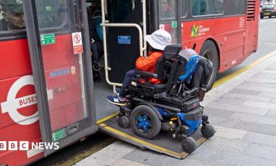 A person in a motorised wheelchair wearing an orange jacket and a white hat boards a bus in London using a wheelchair ramp in October 2025