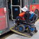 A person in a motorised wheelchair wearing an orange jacket and a white hat boards a bus in London using a wheelchair ramp in October 2025