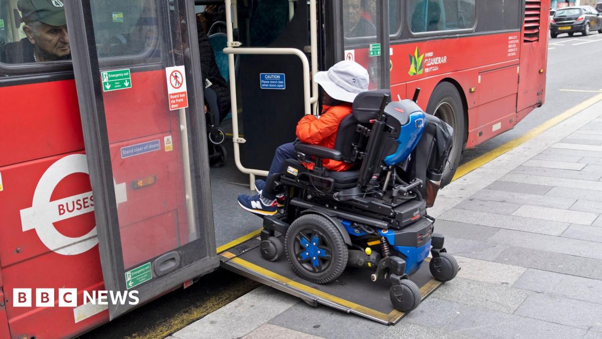 A person in a motorised wheelchair wearing an orange jacket and a white hat boards a bus in London using a wheelchair ramp in October 2025