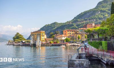 Sunny yellow buildings and greenery sit infront of green mountains overlooking Lake Como.