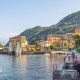 Sunny yellow buildings and greenery sit infront of green mountains overlooking Lake Como.