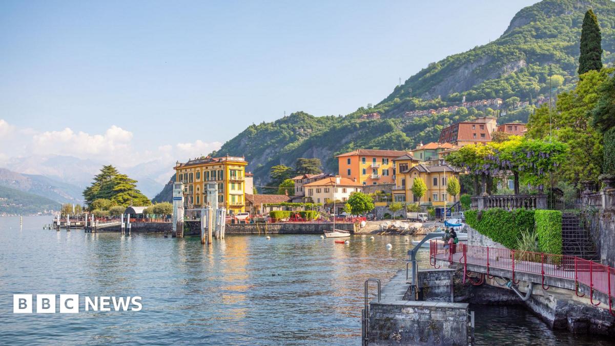 Sunny yellow buildings and greenery sit infront of green mountains overlooking Lake Como.