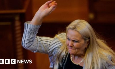 Blond woman wearing white and black blazer lifting right arm and holding egg
