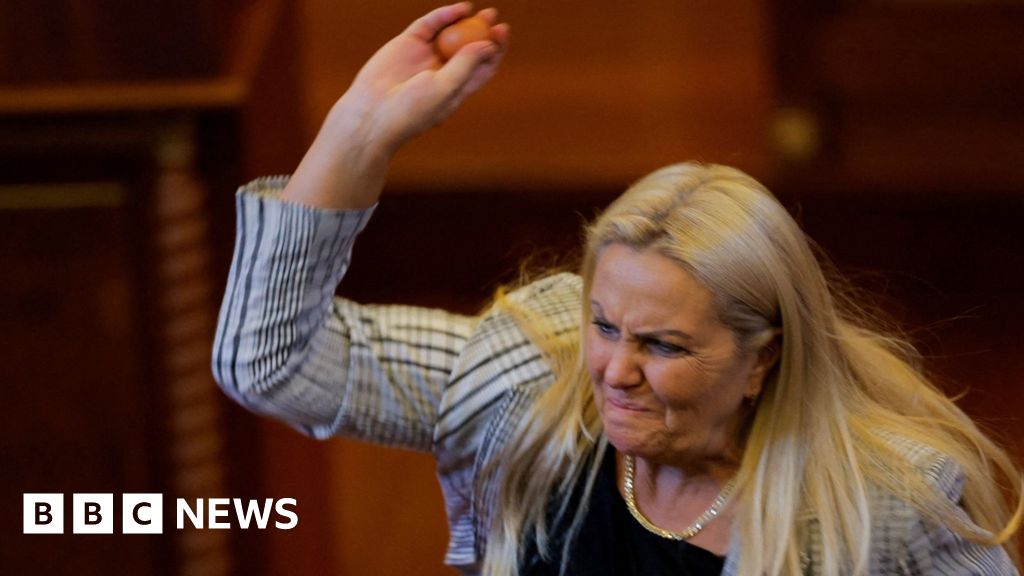 Blond woman wearing white and black blazer lifting right arm and holding egg