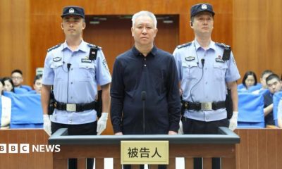 China Evergrande Group founder Hui Ka Yan takes the stand in a court in China. He has a blank look and is standing between two officers, with members of the audience in the background.