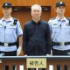 China Evergrande Group founder Hui Ka Yan takes the stand in a court in China. He has a blank look and is standing between two officers, with members of the audience in the background.