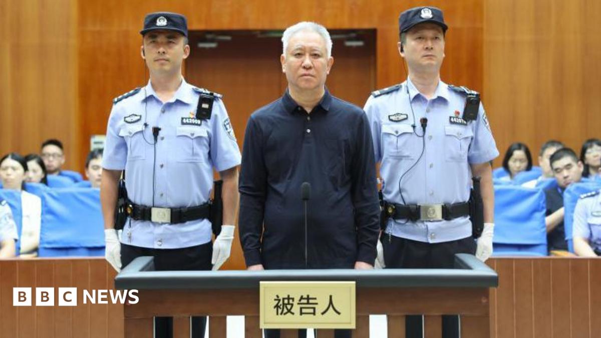 China Evergrande Group founder Hui Ka Yan takes the stand in a court in China. He has a blank look and is standing between two officers, with members of the audience in the background.