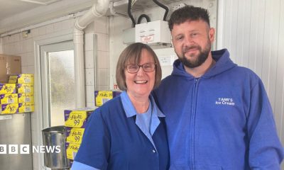 Deana Pimm and Bradley Tarr wearing branded blue tops and overalls standing in the back of the shop arm in arm smiling at the camera.