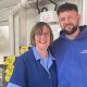 Deana Pimm and Bradley Tarr wearing branded blue tops and overalls standing in the back of the shop arm in arm smiling at the camera.