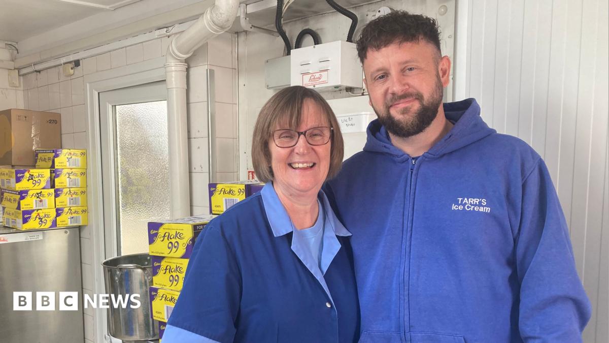 Deana Pimm and Bradley Tarr wearing branded blue tops and overalls standing in the back of the shop arm in arm smiling at the camera.