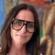 A woman with long dark hair looks at the camera. She is wearing large black glasses