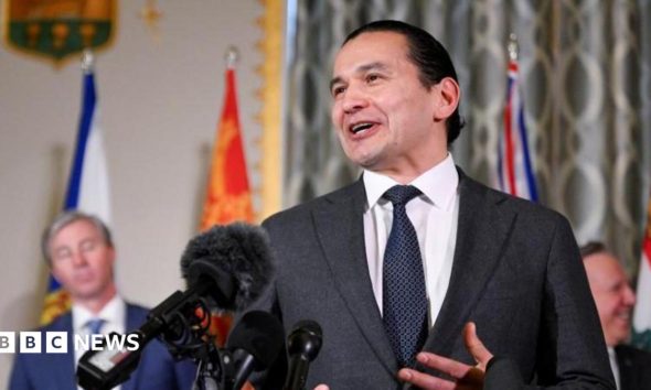 An image showing Wab Kinew speaking in front of several microphones at a press conference with a few of Canada's other premiers behind him. He has black hair that is tied up in a bun, and is wearing a dark charcoal grey suit and tie.