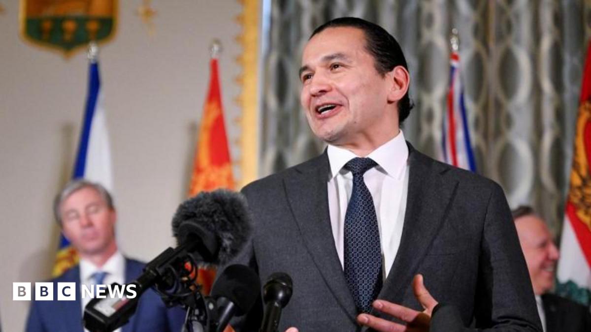 An image showing Wab Kinew speaking in front of several microphones at a press conference with a few of Canada's other premiers behind him. He has black hair that is tied up in a bun, and is wearing a dark charcoal grey suit and tie.