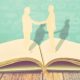 artistic rendering of two people shaking hands and possibly one is a woman building her book of business -- the figures look like cardboard cutouts sitting on top of an open book.