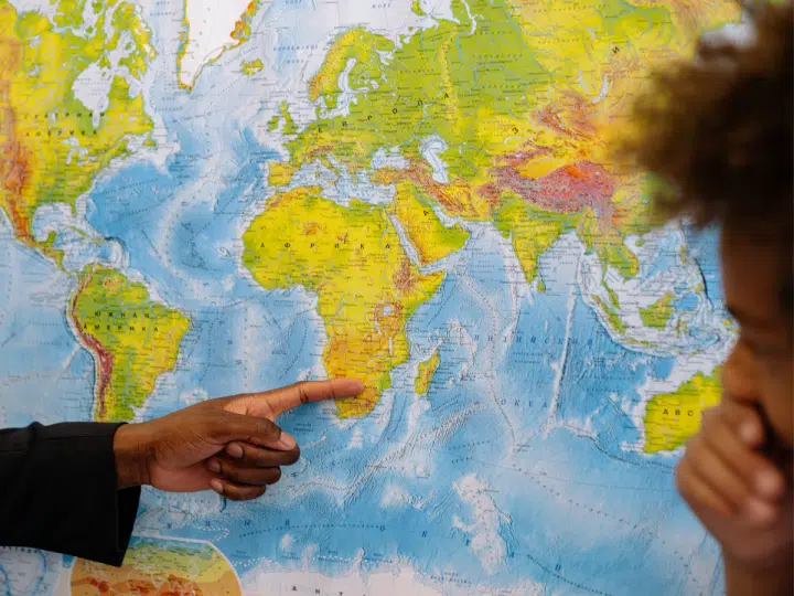 Black male's hand points to Africa on the world map as a black boy looks on from the side with his hand covering his mouth