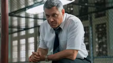 Bill leaning forward in front of a chain link barrier in 'Mindhunter'