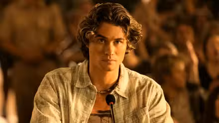 Chase Stokes as John B standing at a podium microphone in Season 4 Episode 6 of Outer Banks