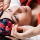 Woman in red satin pajamas lifting a berry-print sleep mask while lying on white bedding