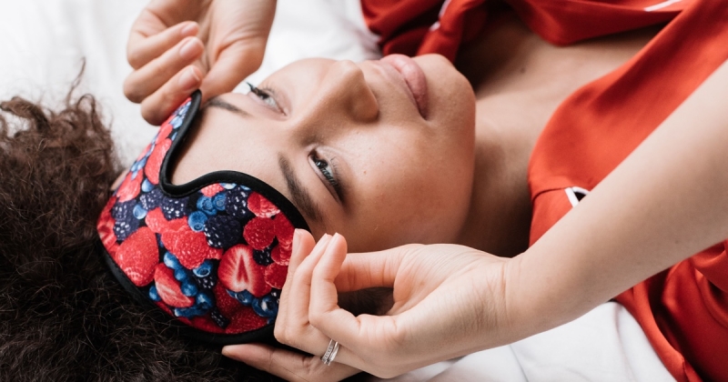 Woman in red satin pajamas lifting a berry-print sleep mask while lying on white bedding