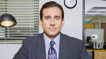 Steve Carell as Michael Scott sitting at a desk in 'The Office'