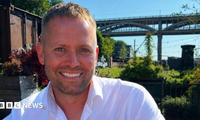 Matthew Blades smiling into the camera. His head and shoulders are visible and he has light brown short hair which is longer on the top. He has short, dark stubble around his mouth and is smiling with his death. He is wearing a white shirt which is open at the top. He is sitting in a garden in front of the High Level Bridge in Newcastle which is a tall, brown structure with arches along the side of it. There are lots of green plants and grass but on the left there is a brown shipping container. There are black fairy lights strung up above the garden and the sky above is blue.