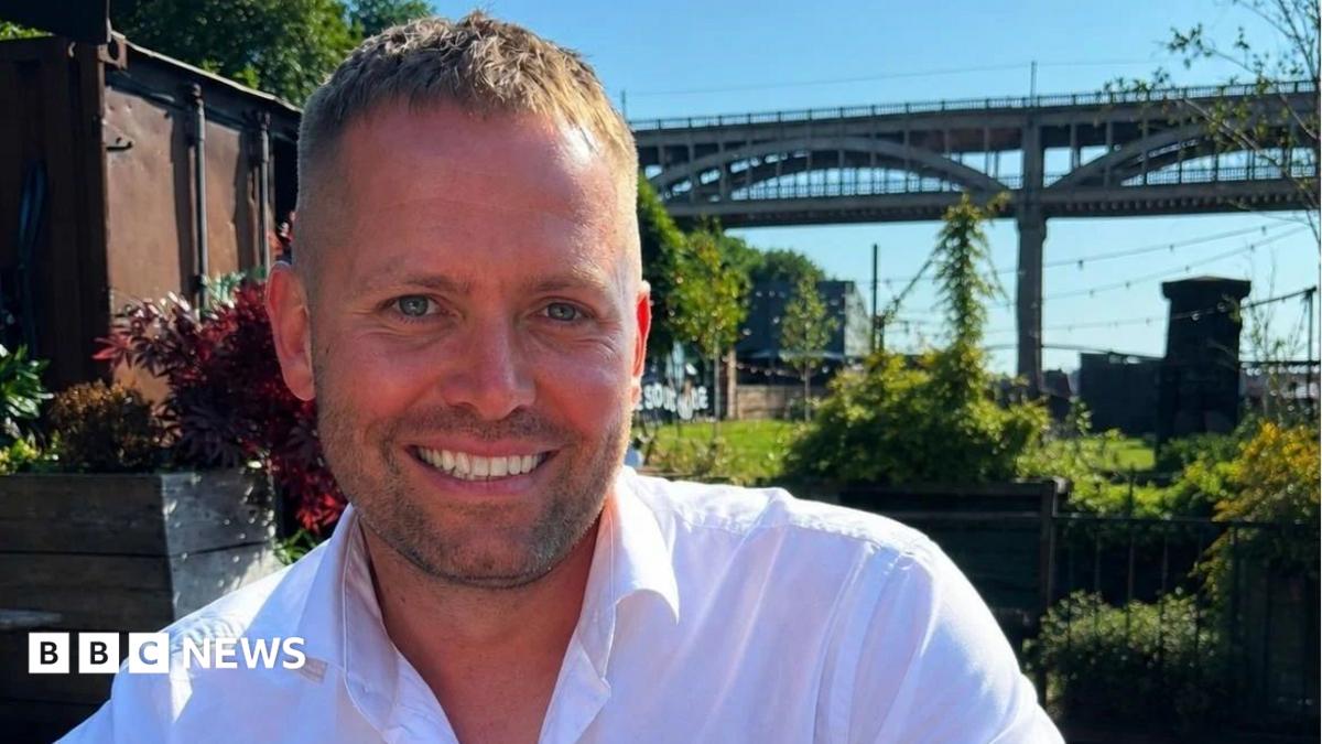 Matthew Blades smiling into the camera. His head and shoulders are visible and he has light brown short hair which is longer on the top. He has short, dark stubble around his mouth and is smiling with his death. He is wearing a white shirt which is open at the top. He is sitting in a garden in front of the High Level Bridge in Newcastle which is a tall, brown structure with arches along the side of it. There are lots of green plants and grass but on the left there is a brown shipping container. There are black fairy lights strung up above the garden and the sky above is blue.