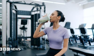 A young woman is sat in a gym drinking a protein shake. She is wearing a purple cropped top and navy blue leggings.