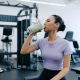 A young woman is sat in a gym drinking a protein shake. She is wearing a purple cropped top and navy blue leggings.