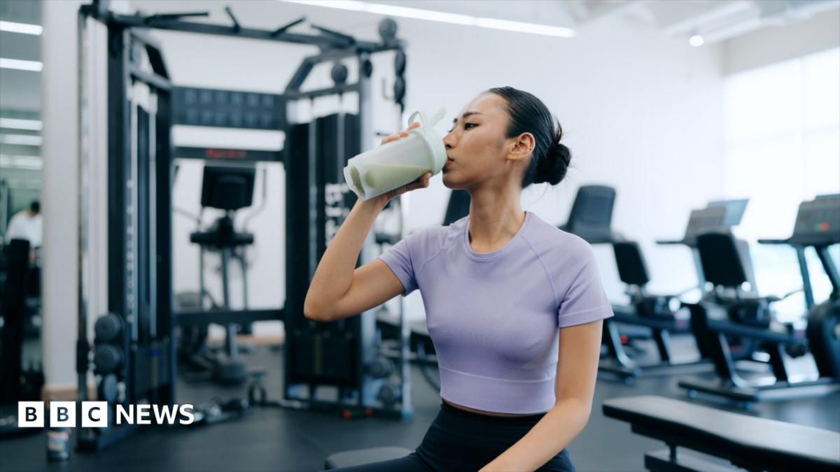 A young woman is sat in a gym drinking a protein shake. She is wearing a purple cropped top and navy blue leggings.