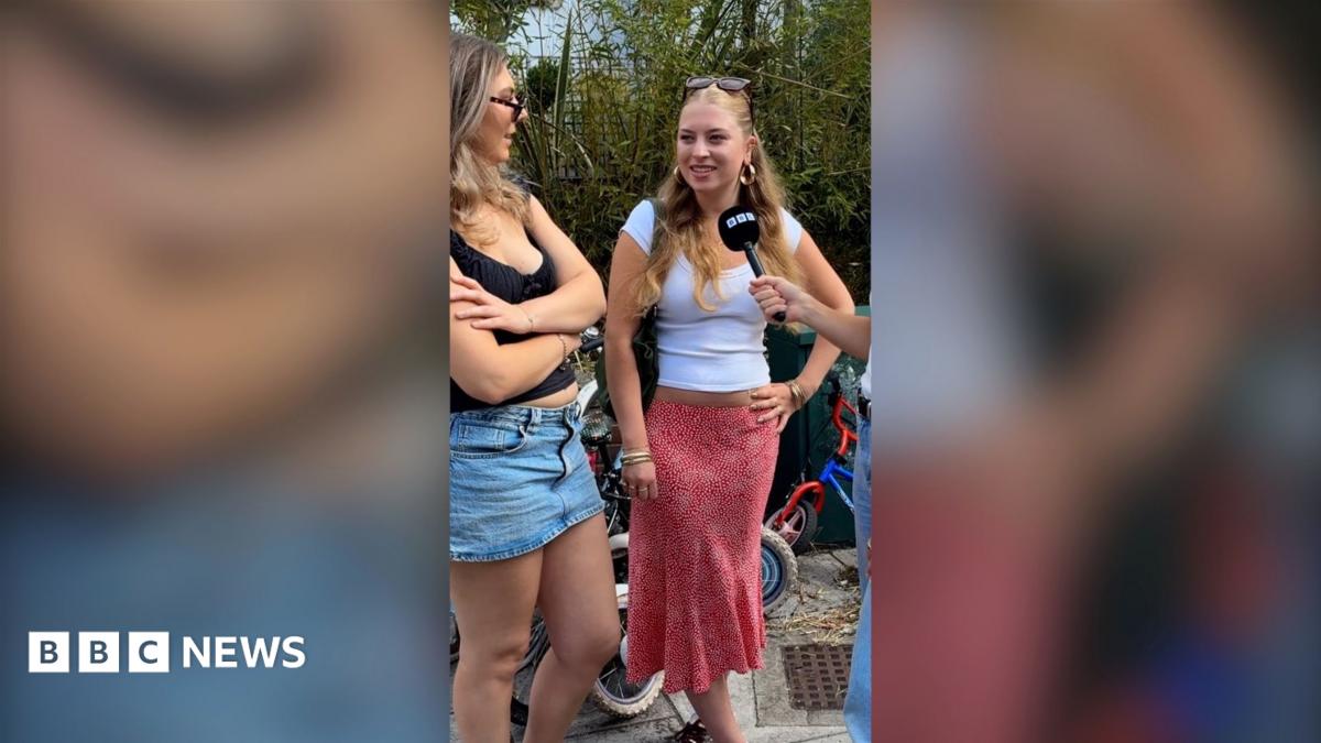 A woman with a white T-shirt and red skirt smiles in front of a mic. She's in the street with her friend.