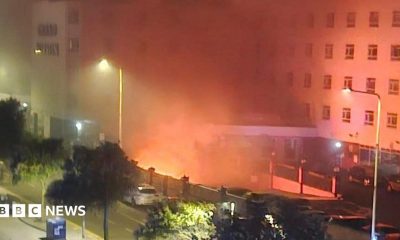 A fire in the basement of a hotel. Flames can be seen from below the building, and emergency services can be seen in the foreground.