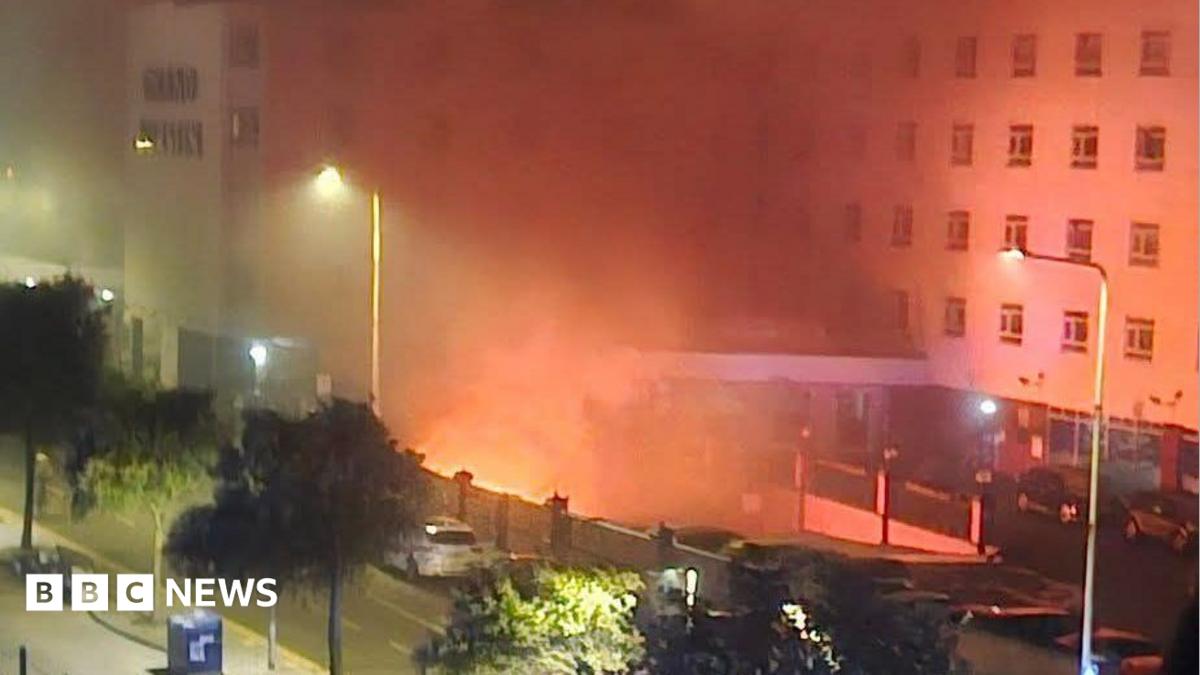 A fire in the basement of a hotel. Flames can be seen from below the building, and emergency services can be seen in the foreground.