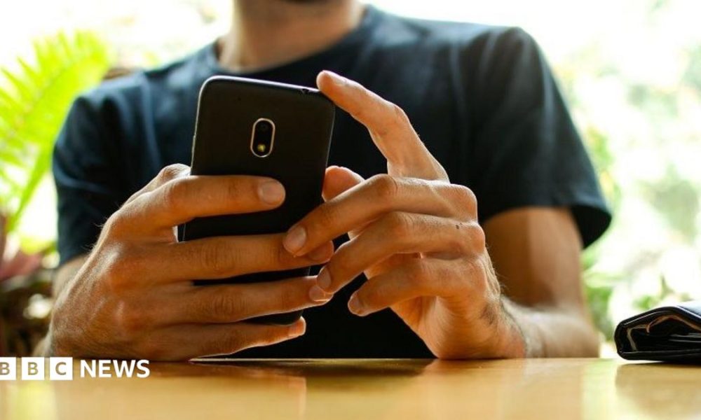 A man, whose face has been cut off in the picture, holding a phone with both hands while resting his arms on a wooden table. There are out-of-focus plants in the background. He's wearing a dark green t shirt.