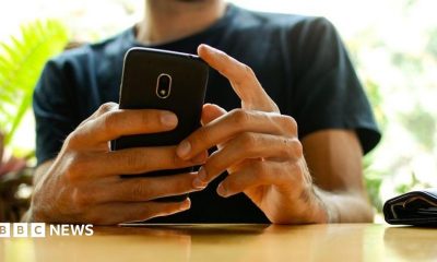 A man, whose face has been cut off in the picture, holding a phone with both hands while resting his arms on a wooden table. There are out-of-focus plants in the background. He's wearing a dark green t shirt.