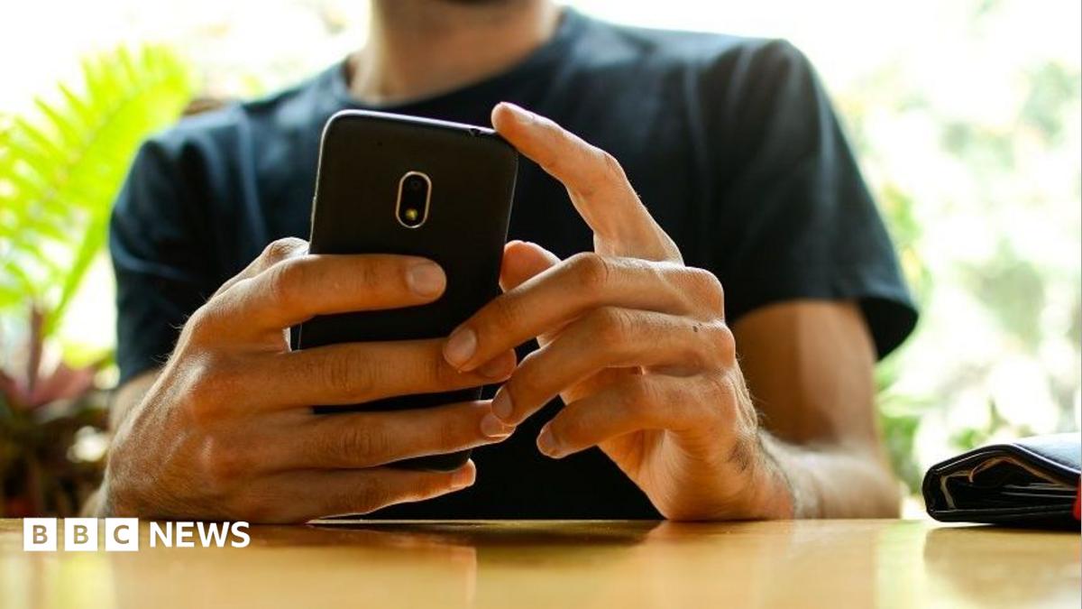 A man, whose face has been cut off in the picture, holding a phone with both hands while resting his arms on a wooden table. There are out-of-focus plants in the background. He's wearing a dark green t shirt.