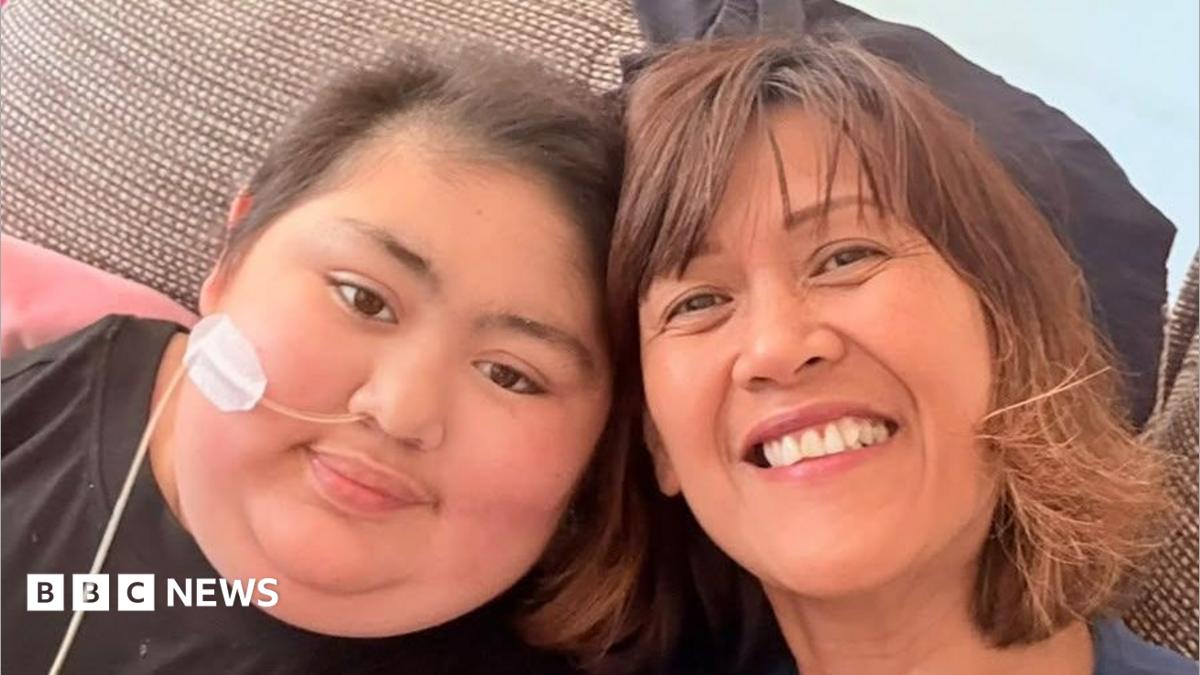 A woman and her daughter, sitting next to each other and smiling into a camera. The daughter, left, has short hair and has a tube inserted into her nose, which is taped to her face.