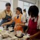 Two teenagers at a classroom bench, wearing aprons., They are in a catering class and are adding cream cheese to bowls, and are being supervised by their female teacher, also in an apron, who is standing to the left of them.