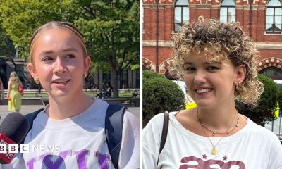 Composite image of two women. One woman has blonde hair tied back and wears a grey sweater; she is standing in a park speaking into a mic. The other woman has curly blonde hair and smiles standing in the street; she wears a white T-shirt.