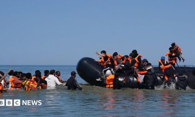 People thought to be migrants attempt to board a small boat off the coast of Hardelot, France, during an attempt to cross the English Channel on 29 July 2026. A group of people are on the inflatable vessel wearing orange life jackets and others are in the water, waiting to board the dinghy.