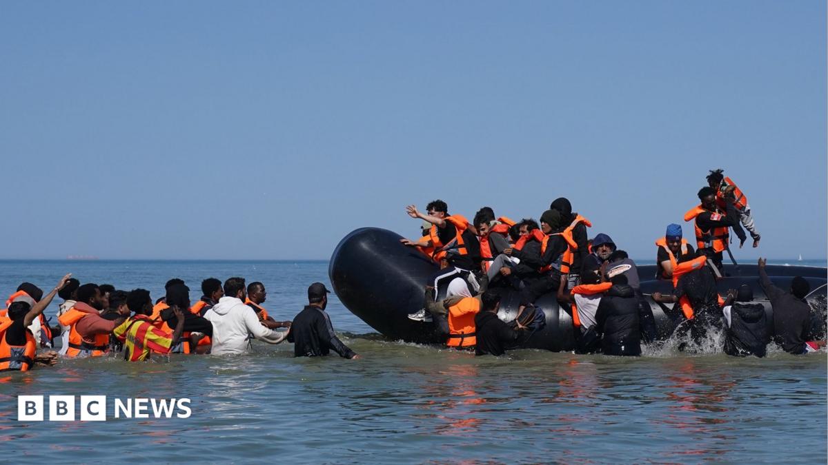 People thought to be migrants attempt to board a small boat off the coast of Hardelot, France, during an attempt to cross the English Channel on 29 July 2026. A group of people are on the inflatable vessel wearing orange life jackets and others are in the water, waiting to board the dinghy.
